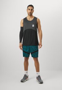 Débardeur sportif sans manches noir et gris avec un patch logo, associé à un short teal avec un motif léopard noir. Chaussures de sport grises.