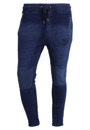 Granatowe, zwężane spodnie dresowe z prążkowanymi detalami na kolanach, zamykanymi na zamek kieszeniami po bokach, elastycznym pasem i czarnymi sznurkami do ściągania.