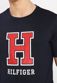 Mörkblå bomulls-t-shirt med en stor röd "H"-logotyp med vit kontur, och texten "HILFIGER" i vitt nedanför. Kortärmad, rund halsringning.