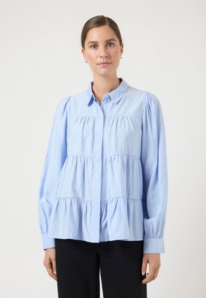 Vrouw met haar in een staart draagt een lichtblouse blouse met lange pofmouwen en een zwarte broek tegen een effen achtergrond.