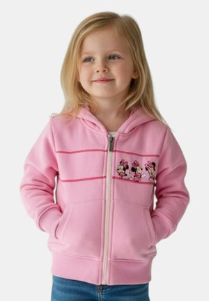Ragazza giovane con capelli biondi che indossa una felpa con cappuccio rosa con cerniera e motivi di Minnie Mouse e jeans blu, mani in tasca, sorride leggermente.