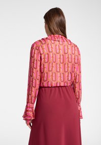 Vrouw met lang bruin haar, gekleed in een roze blouse met patroon en een hooggesneden effen bordeauxrode rok, gezien van achteren.