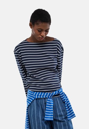 Femme portant un haut à manches longues à rayures bleu marine et blanches, un pull bleu rayé noué à la taille, et un pantalon bleu à fines rayures, regardant vers le bas.