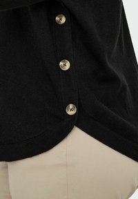 Cardigan nero in maglia con quattro bottoni marrone chiaro sopra pantaloni beige, che mostra la cucitura laterale e l'orlo curvo del cardigan.