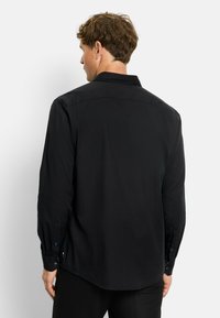 Mann mit kurzen braunen Haaren, der ein schwarzes Langarmshirt trägt, von hinten vor einem schlichten hellen Hintergrund dargestellt.