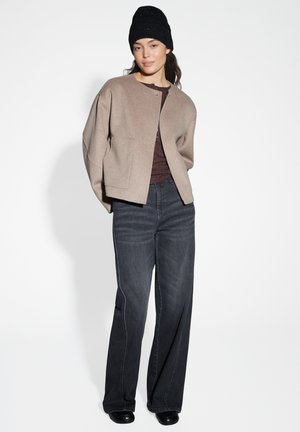 Beige Wollmischjacke mit runder Halslinie, kombiniert mit dunkelblauen, weit geschnittenen Jeans. Eine schwarze Strickmütze sorgt für Textur.