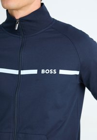 Sötétkék, cipzáros dzseki sima anyagból, fehér vízszintes csíkkal, a "BOSS" márkanevével hangsúlyosan a mellkason.