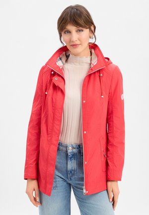 Vrouw met lichtbruin haar, gekleed in een rode jas met capuchon over een beige geribbelde trui en lichtblauwe spijkerbroek, staand tegen een effen achtergrond.