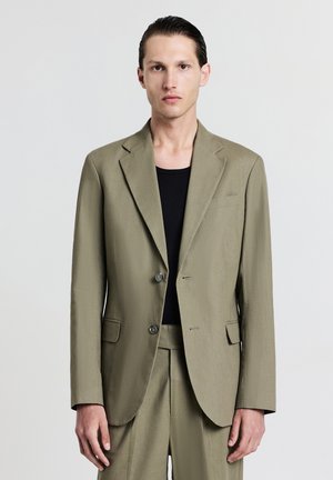 Blazer vert olive en tissu lisse, doté de deux boutons, d'un revers cranté et de deux poches avant. Associe à un haut noir en dessous.