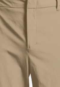 Primo piano dei pantaloni beige frontalmente, che mostrano un tessuto liscio con cuciture visibili e una passante per cintura parzialmente visibile.