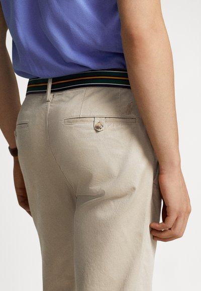 Osoba ubrana w beżowe spodnie z tylnymi kieszeniami zapinanymi na guziki, paskiem w paski i włożoną do środka niebieską koszulę, widoczna od talii w dół.