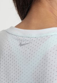 Vaaleansininen urheilupaita, jossa on hengittävä verkkotekstuuri, ribattu pyöreä kaula-aukko ja pieni harmaa Nike-logo takana.