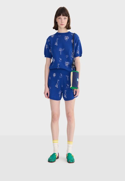 Kobieta w niebieskim, dopasowanym topie z krótkim rękawem i szortach z białymi wzorami ptaków i roślin, niosąca małą, granatowo-zieloną torbę na ramię.