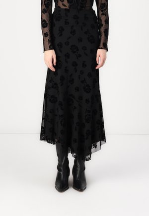 Femme portant une jupe longue noire fleurie transparente avec un ourlet asymétrique et des bottes noires à bout pointu, debout devant un fond uni.