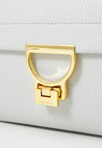 Sac à main en cuir texturé blanc avec une poignée circulaire en métal doré et un fermoir doré portant le nom de la marque "Coccinelle" gravé.