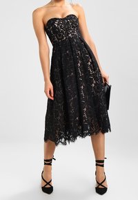 Mustat pitsiset olkaimettomat mekko, jossa on tiukka yläosa ja leveä helma, ja jossa on aaltoileva helman reuna. Yhdistetty mustiin remmillisiin korkokenkiin.