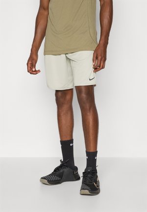 Mann trägt beige Nike-Shorts, schwarze Nike-Socken und schwarze Nike-Sneaker und steht vor einem schlichten hellen Hintergrund.