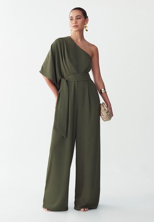 Donna che indossa una tuta monospalla verde oliva, tiene una clutch intrecciata, con grandi orecchini a forma di foglia dorati e un bracciale rigido dorato.