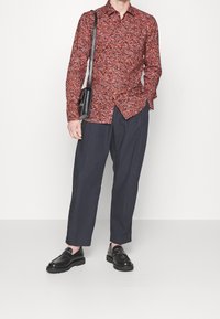 Camicia a bottoni con motivo floreale rosso, pantaloni blu navy larghi e mocassini neri con una leggera piattaforma, abbinati a una borsa nera.