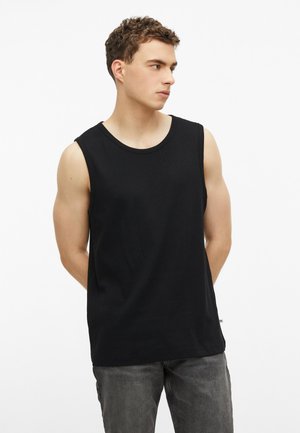 Junger Mann mit kurzen lockigen Haaren, der ein schwarzes ärmelloses Shirt und graue Jeans trägt, schaut seitlich mit den Händen hinter dem Rücken.