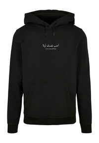 Fekete kapucnis pulóver, puha anyagból, elülső zsebbel és fehér szöveggel arab és angol nyelven: "Szeresd először önmagad."