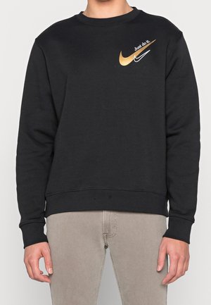 Person trägt schwarzen Pullover mit goldenen und weißen Nike-Logos und dem Schriftzug „Just do it.“, kombiniert mit beigen Hosen, steht vor weißem Hintergrund.