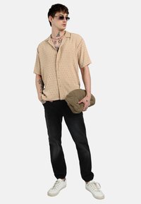 Camisa beige de manga corta con patrón texturizado, pantalones negros ajustados y zapatillas blancas. El modelo sostiene una chaqueta verde acolchada.