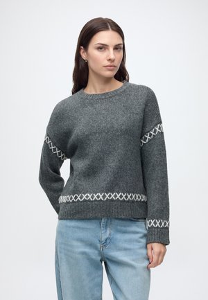 Femme portant un pull gris foncé avec des motifs blancs en croix sur les manches et la taille, associée à un jean bleu clair, debout devant un fond uni.