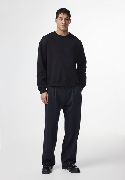 Schwarzer Sweatshirt mit einem schlichten Design, ausgestattet mit einem runden Ausschnitt und gerippten Bündchen. Kombiniert mit dunklen, plissierten Hosen und schwarzen Schuhen.