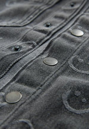 Sort denim stof med metalknapper, prydet med en broderet smiley ansigt mønster. Glat tekstur og tæt fokus på detaljerne.