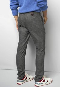 Mann in grauen maßgeschneiderten Hosen, weißen Sneakers mit roten und blauen Details und einem blauen Strickpullover, der vor einem schlichten Hintergrund steht.