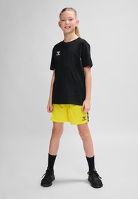 Schwarzes T-Shirt mit kurzen Ärmeln und Logo, kombiniert mit leuchtend gelben Shorts mit seitlichen Streifen und schwarzen Akzenten. Schwarze Sportschuhe und Socken.