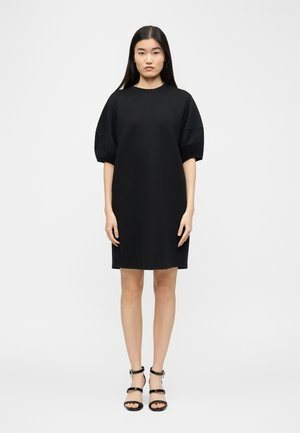 Femme aux longs cheveux noirs portant une robe noire ample jusqu'au genou avec des manches bouffantes et des talons noirs à brides, debout devant un fond uni.