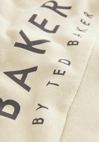 Platno u krem boji s velikim crnim tekstom koji kaže "BAKER" i manjim tekstom "by TED BAKER", s teksturiranom površinom.