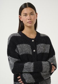 Knitted cardigan featuring large alternating black and gray check patterns. It has a round neck, button closure, and ribbed cuffs. Soft texture.