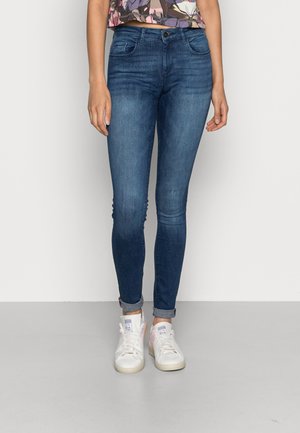 Femme portant un jean slim bleu marine retroussé aux chevilles et des baskets blanches avec un haut court coloré.