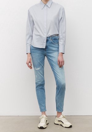 Chemise bleu clair boutonnée, ajustée avec manches longues. Portée avec un jean bleu clair déchiré et des baskets beiges avec des accents noirs.