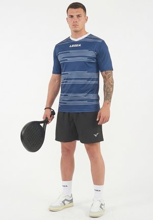 Atleta con maglia blu a motivi e pantaloncini neri impugna una pagaia nera, in piedi su sfondo bianco, indossa calzini bianchi e scarpe da ginnastica.