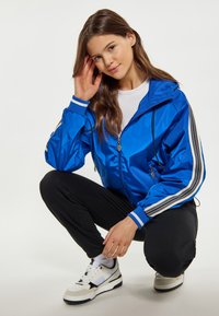 Giacca leggera blu con cappuccio, chiusura con zip, strisce bianche e grigie a contrasto sulle maniche, indossata sopra una t-shirt bianca e pantaloni neri.