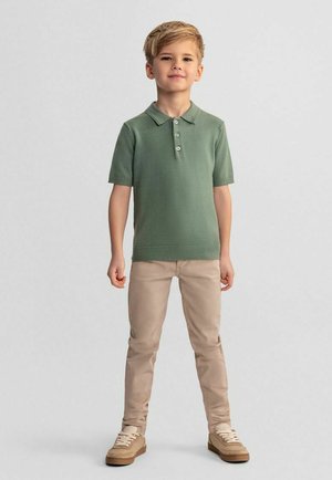 Chłopiec stojący pewnie, ubrany w zieloną, krótką koszulkę polo, beżowe spodnie i beżowe trampki na jednolitym szarym tle.