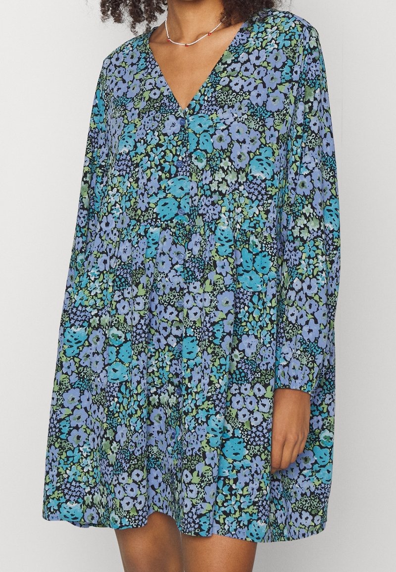 Femme portant une robe à manches longues, col en V, imprimé floral bleu, vert et noir avec des boutons sur le devant.