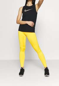 Top atletico senza maniche nero con logo Nike riflettente, abbinato a leggings gialli attillati e scarpe da ginnastica nere.