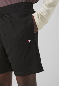 Sort meshshorts med elastisk taljebånd, der har sidelommer og et lille broderet logo på venstre ben.