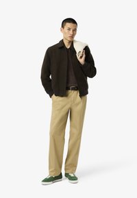 Nuorisokaveri seisoo itsevarmana, päällään tumma takki, ruskea paita, vaaleat housut ja vihreät lenkkarit, pitäen kermankeltaista villapaitaa olkapäällään.