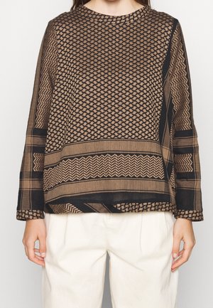 Mujer con un top marrón y negro de manga larga con diseños geométricos y pantalones blancos, de pie frente a un fondo liso.