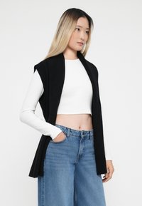 Musta hihaton cardigan ribbetekstuuri kanssa, kerrottu valkoisen crop-topin päällä, yhdistetty korkeavyötäröisten sinisten farkkujen kanssa.