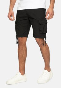 Schwarze Cargo-Shorts mit mehreren Seitentaschen, Kordelzugsäumen und einer lockeren Passform, kombiniert mit weißen Sneakers. Aus Baumwollmaterial.