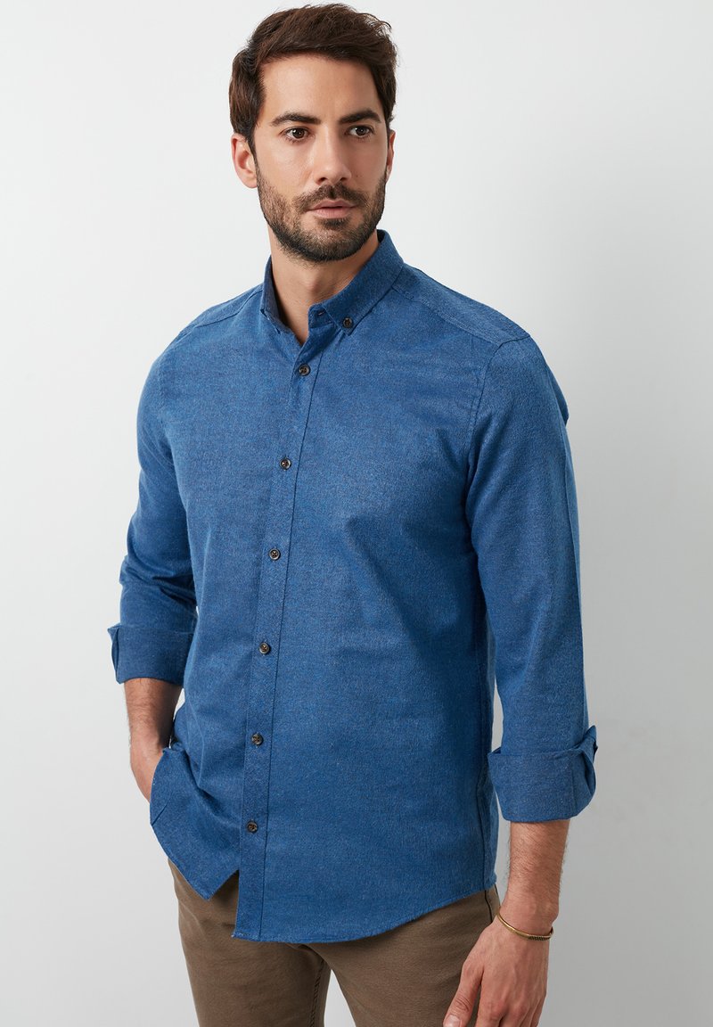 Camisa azul de botones en una tela texturizada con cuello y mangas remangadas, que presenta botones oscuros y un dobladillo curvado.