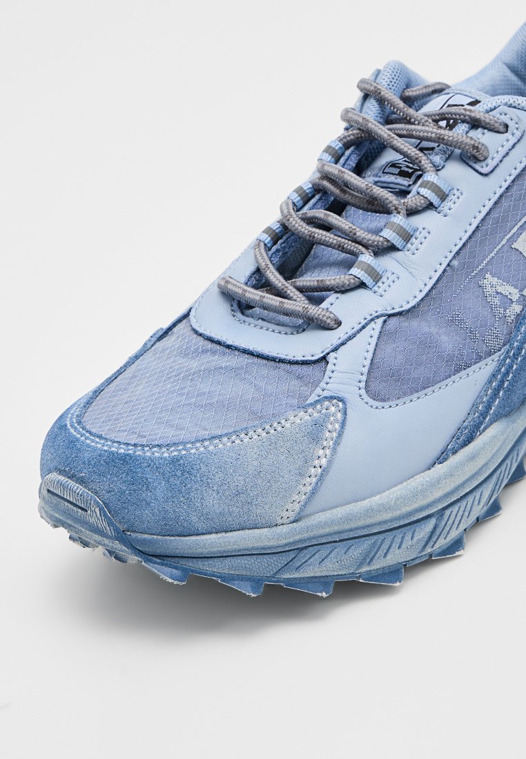 Zapatilla deportiva de color azul claro con paneles de ante y malla, costuras detalladas, cordones grises y suela de goma texturizada sobre un fondo blanco.