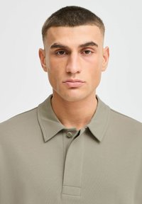 Khaki värvi polo särk klassikalise kraega, nööbiga kinnitusega ja lühikeste varrukatega, valmistatud siledast puuvillakangasest ning sobib keha lähedale.